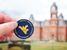 A hand holds a WVU values coin in front of woodburn hall