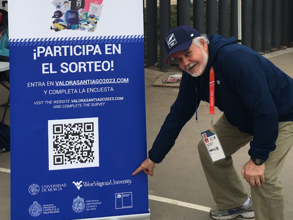
              Gonzalo Bravo stands next to a sign at the Pan American Games and points to the West Virginia University logo.
            
