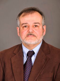 Dr. Bravo has grey hair, beard and mustache and is wearing a dark brown dress jacket, light blue dress shirt, dark blue tie with dots.